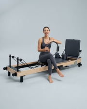 Femme souriante assise sur le reformer Pilates pliable Opilat coloris Charbon, avec un anneau de Pilates