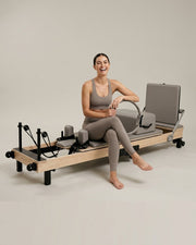 Femme souriante assise sur le reformer Pilates pliable Opilat coloris Galet, avec un anneau de Pilates