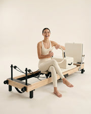 Femme souriante assise sur le reformer Pilates pliable Opilat coloris Ivoire, avec un anneau de Pilates